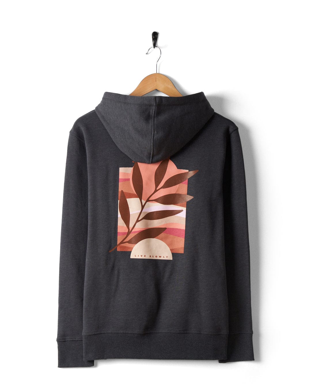 The Live Slowly - Women's Pop Hoodie in grey, by Saltrock, is displayed on a wooden hanger. It features branding and a graphic of abstract leaves and shapes in brown, white, and pink on the back.