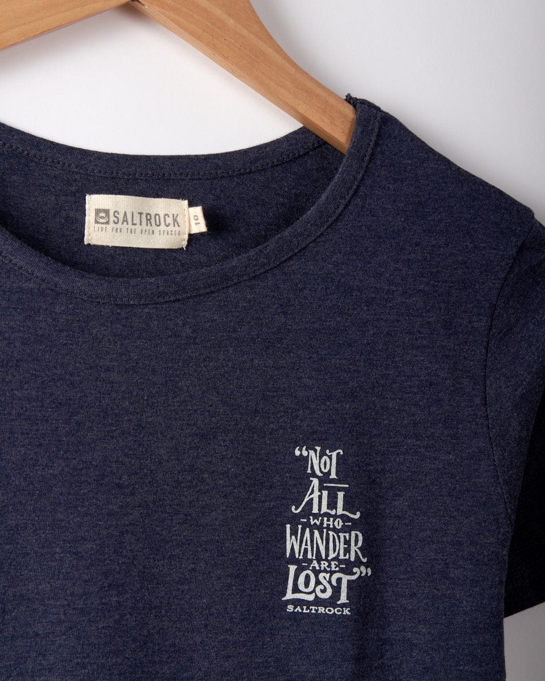 Close-up of the neckline of a dark blue Saltrock Lost Ships women's t-shirt on a wooden hanger. The shirt, made from 100% cotton and machine washable, features the text: "Not all who wander are lost" in white.