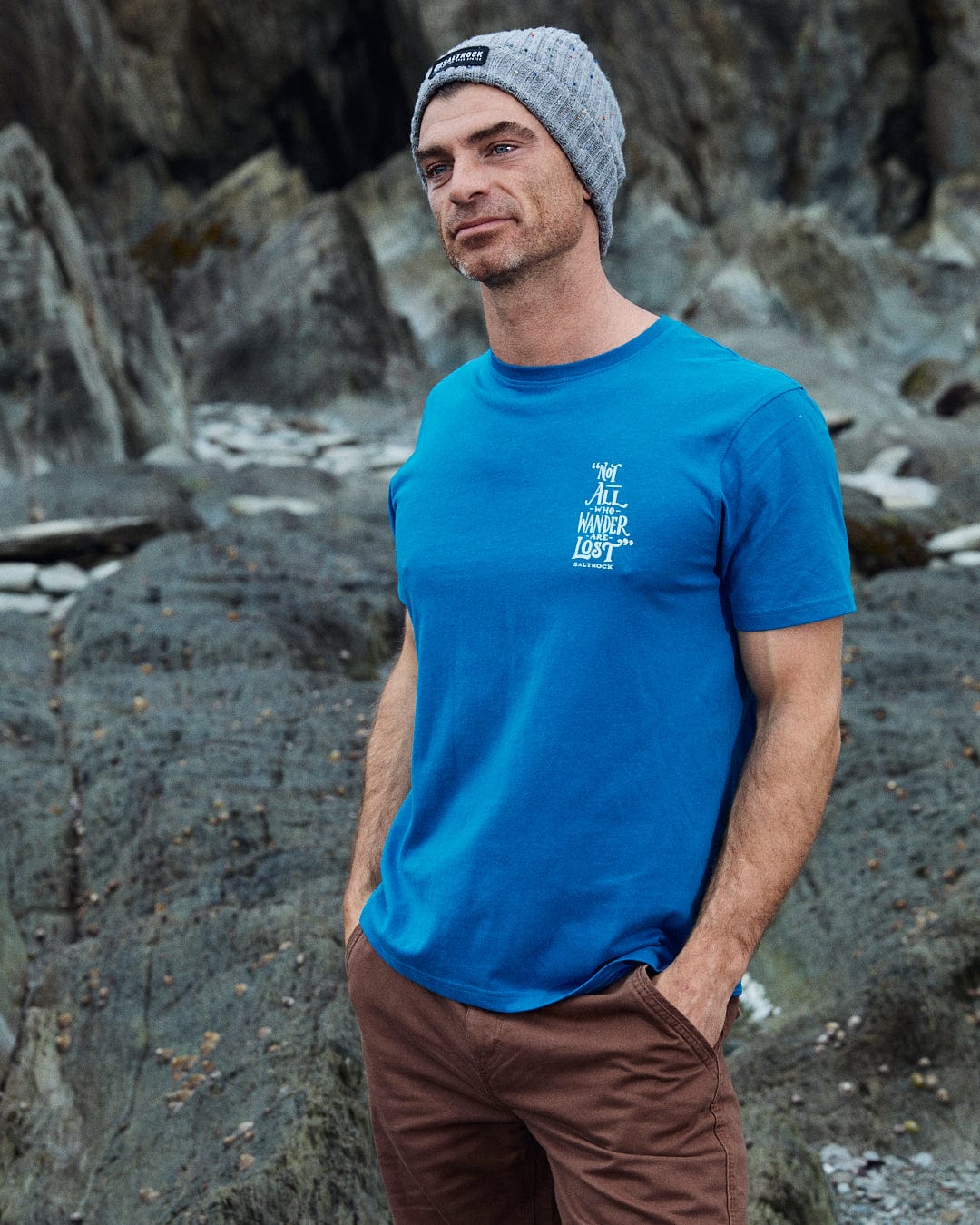 A man wearing a Saltrock classic gray beanie, blue Lost Ships men's short sleeve T-shirt, and brown pants stands with hands in pockets on rocky terrain, gazing into the distance.