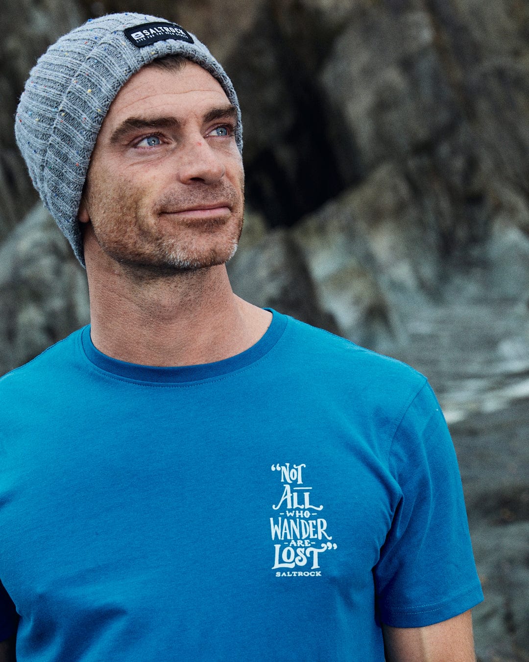 A man in a blue "Lost Ships" T-shirt by Saltrock and a gray beanie stands against rocky terrain, highlighting Saltrock's classic 100% cotton style.