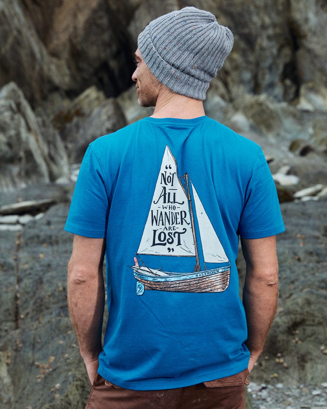 A man in a Saltrock Lost Ships blue T-shirt, made of 100% cotton with "Not all who wander are lost" and sailboat graphic, stands outdoors in rocky terrain wearing a gray beanie.