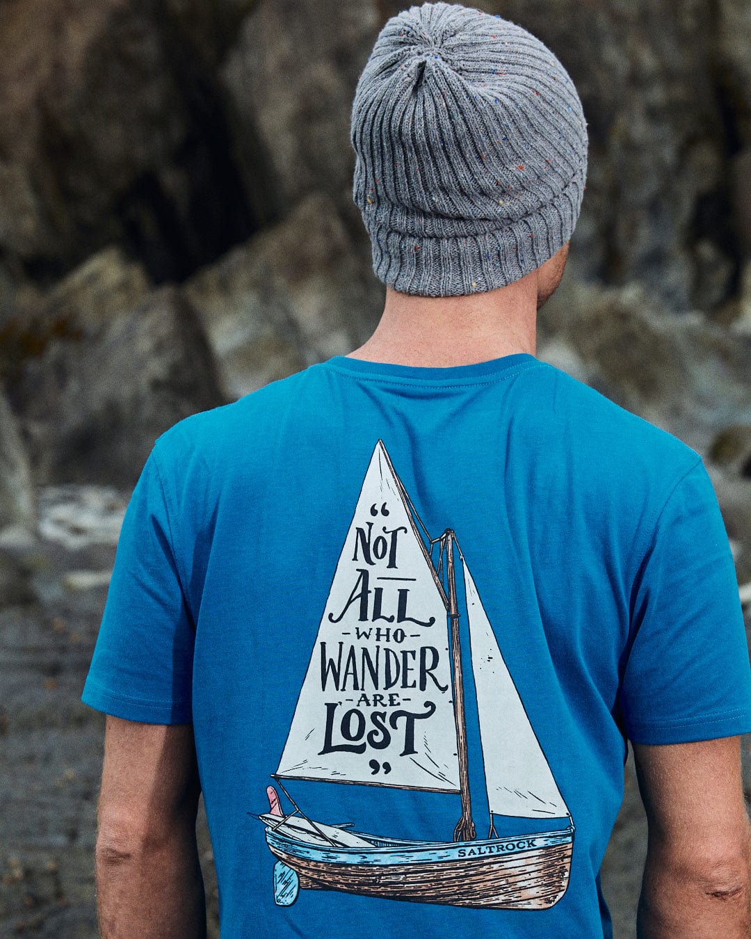 A person in a gray beanie and the Saltrock Lost Ships Men's Short Sleeve T-Shirt in blue, featuring a sailboat design and "Not all who wander are lost," showcases classic Saltrock style outdoors.