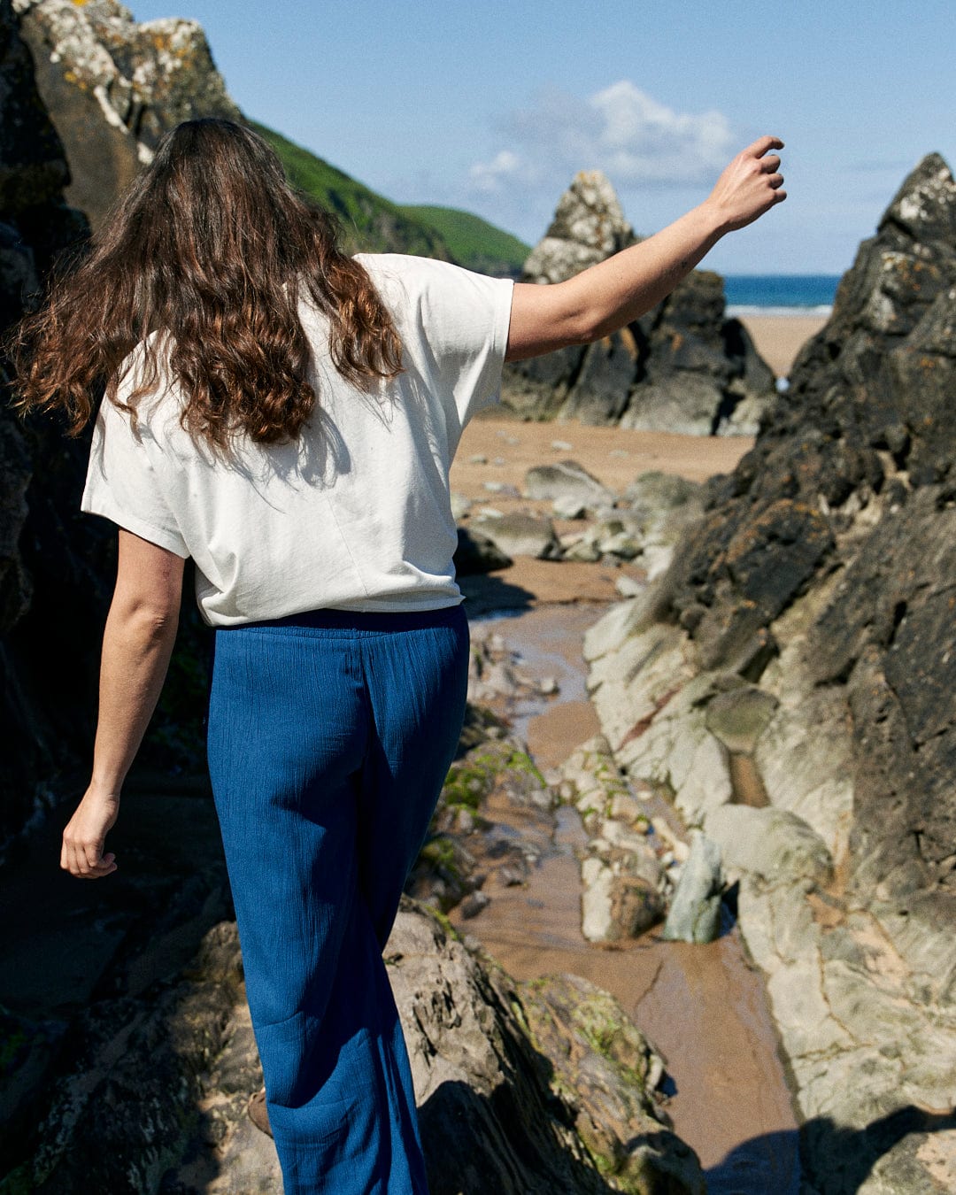 A person with long hair, clad in a loose fit white Saltrock Love Your Ocean - Recycled Womens T-Shirt - White and blue pants made from recycled material, is seen from behind while walking on rocks near a sandy beach with green hills in the background. A reminder to "Love Your Ocean" drifts in the salty breeze.