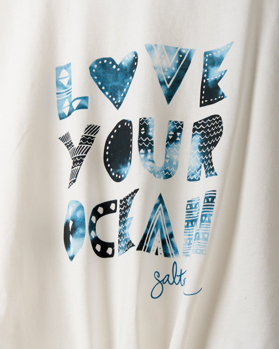 White fabric with the words "LOVE YOUR OCEAN" printed in various blue patterned letters. The word "Saltrock" is written at the bottom in blue script. Made from recycled material, this loose fit tee offers both comfort and a message of sustainability. This is the Love Your Ocean - Recycled Womens T-Shirt - White by Saltrock.