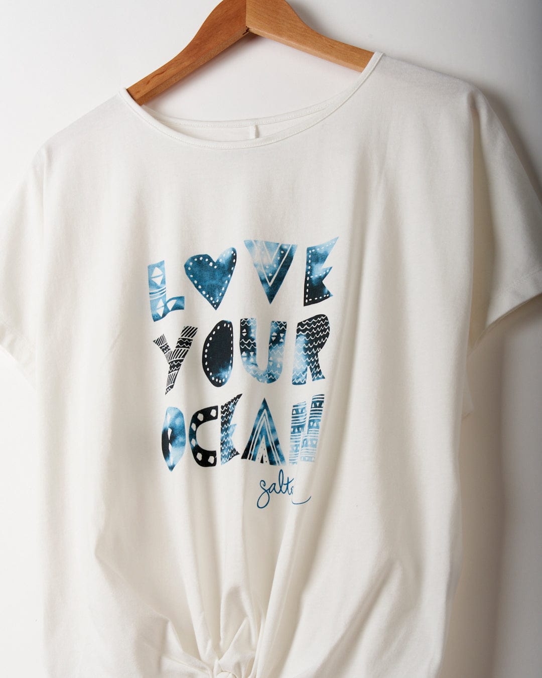 A white, loose fit T-shirt on a wooden hanger with "Love Your Ocean" written in blue patterned letters, featuring a small signature reading "Salt" at the bottom, made from recycled material. The product is the Love Your Ocean - Recycled Womens T-Shirt - White by Saltrock.