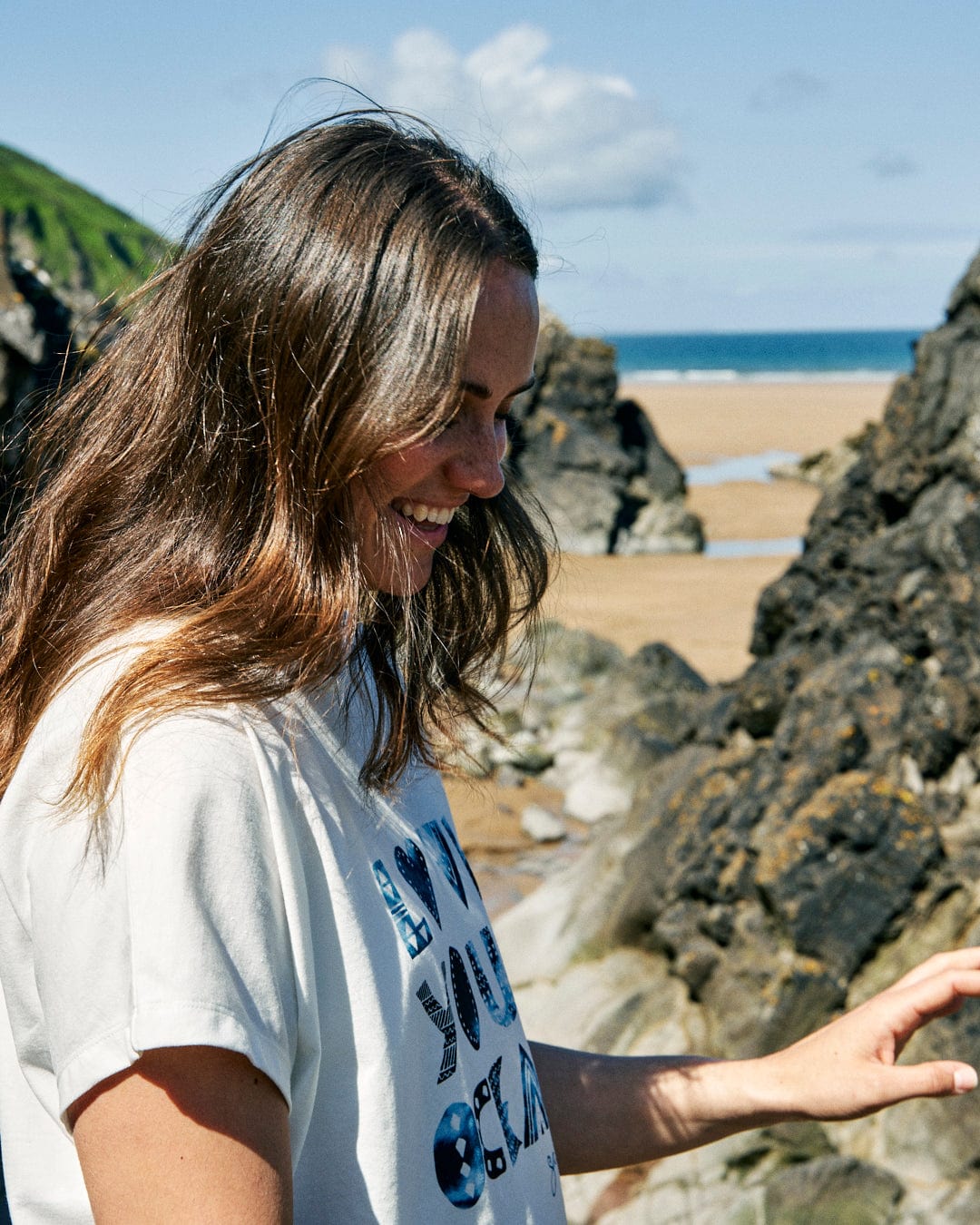 A person with long hair smiles while standing on a rocky beach on a sunny day. They wear a loose fit outfit, perfect for the breezy weather, made from recycled material. In the background, the ocean and sandy shore remind us to Love Your Ocean - Recycled Womens T-Shirt - White by Saltrock.