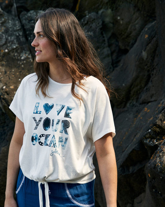 A woman stands outdoors, wearing a loose-fit white Saltrock Love Your Ocean - Recycled Womens T-Shirt - White. She is looking to the side, with dark, wavy hair. Rocks are visible in the background.