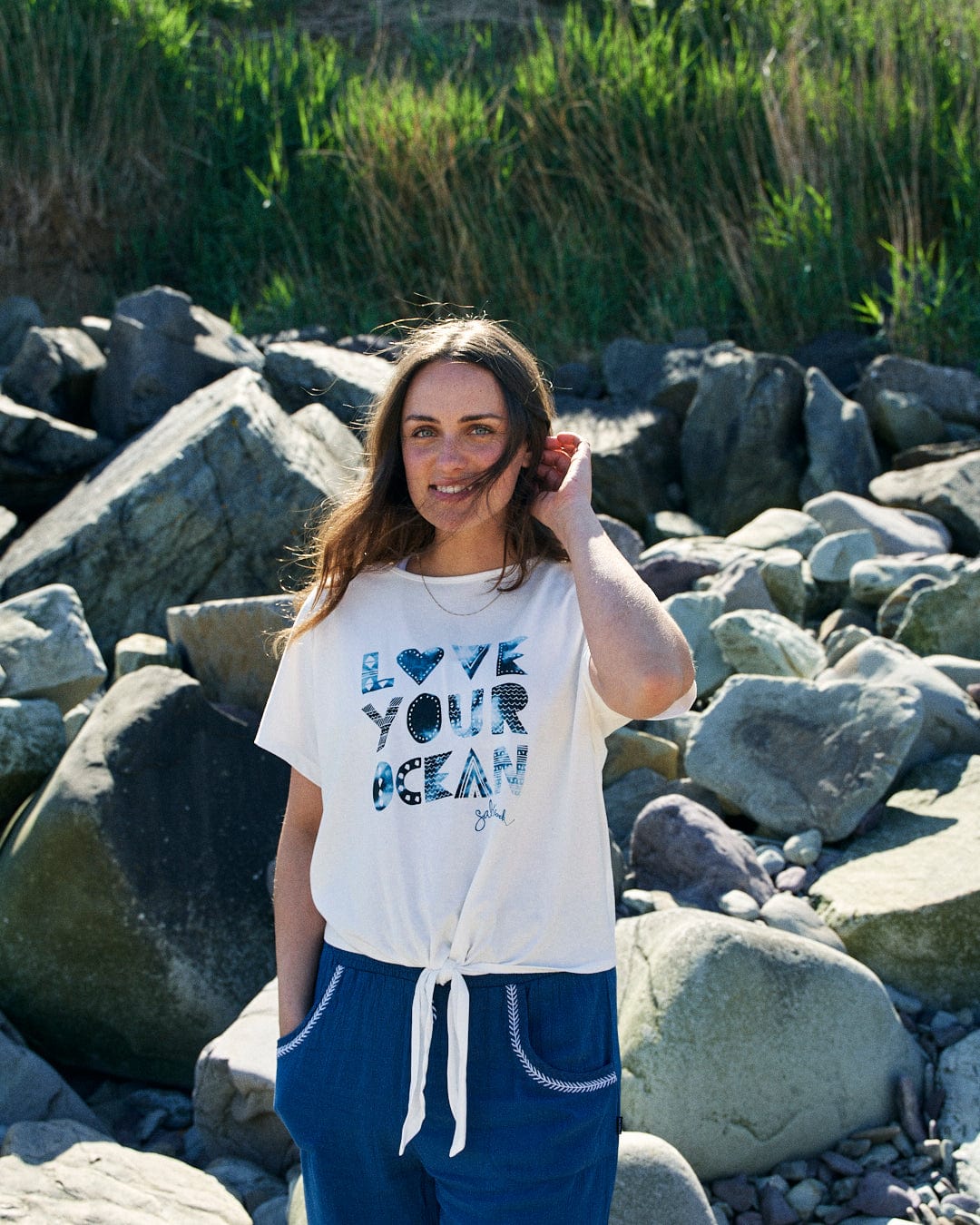 A person with long hair stands on a rocky shoreline wearing a loose-fit white t-shirt that reads "LOVE YOUR OCEAN" and blue pants. The shirt, named **Saltrock's Love Your Ocean - Recycled Womens T-Shirt - White**, is made from recycled material. They are smiling and touching their hair. Grass and rocks are visible in the background.