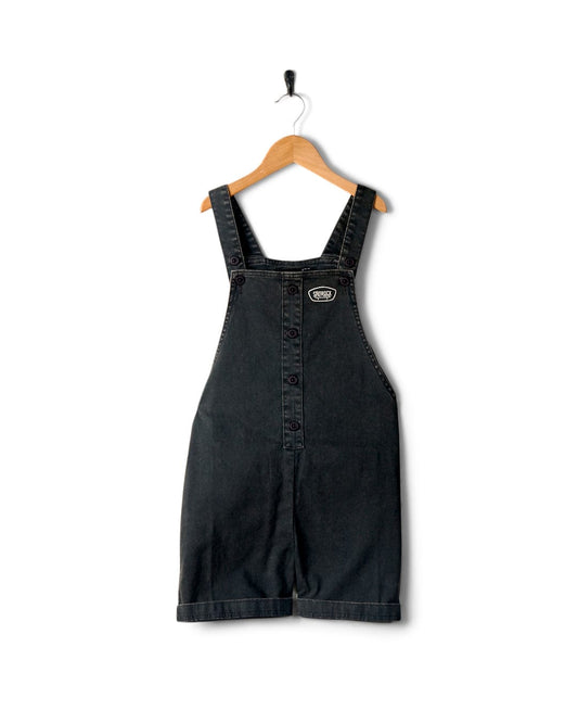 Lyn-Z - Kids Dungarees - Washed Black with adjustable straps hanging on a wooden hanger against a white background, featuring a Saltrock skateboard badge.
