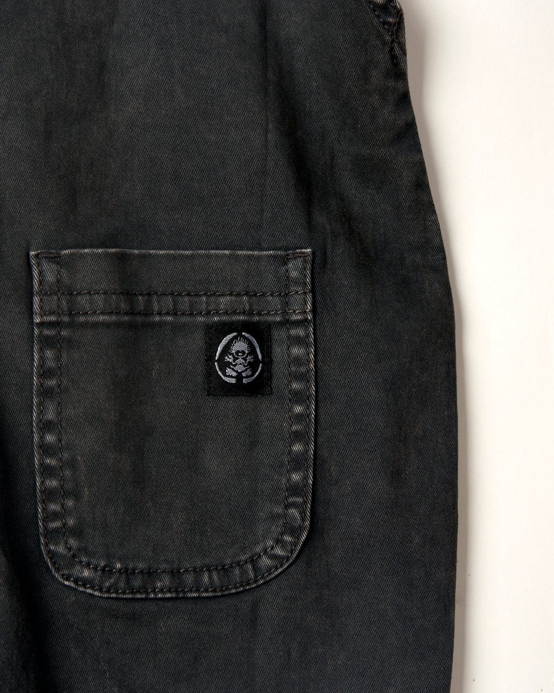 Close-up of an acid wash denim pocket with a stitched label featuring a skull design, part of the Lyn-Z - Kids Dungarees - Washed Black collection by Saltrock.