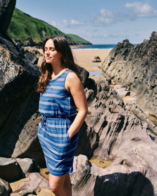 A woman in a knee-length, blue striped Saltrock Marina Bauhaus - Womens Dress - Blue with an elasticated waist stands among large rocks on a sunny beach, green hills rising in the background.