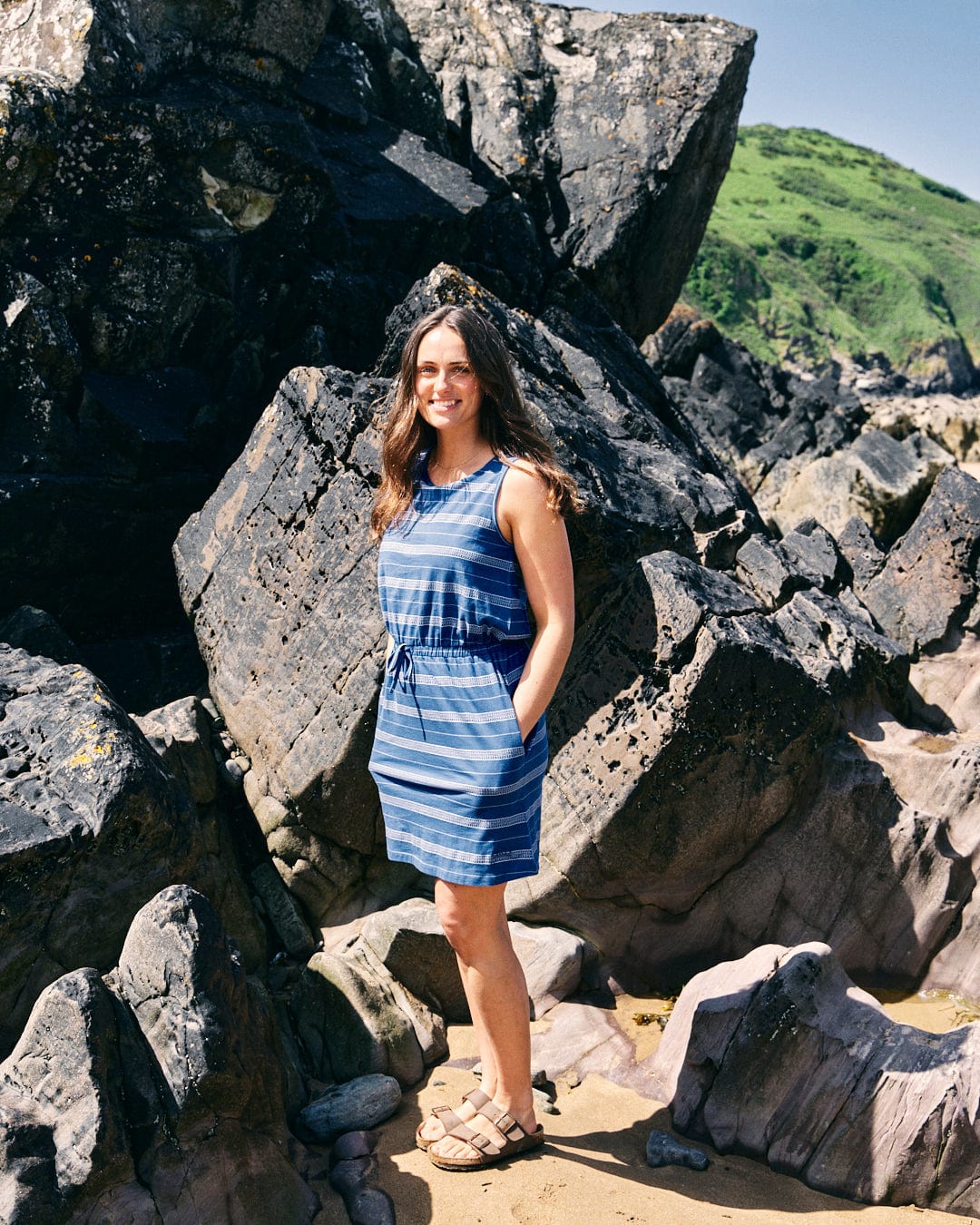 A woman in a Saltrock Marina Bauhaus - Womens Dress - Blue stands smiling in front of large rocks on a sunny day. She is wearing sandals and has her hands in her dress pockets. Green hills are visible in the background.