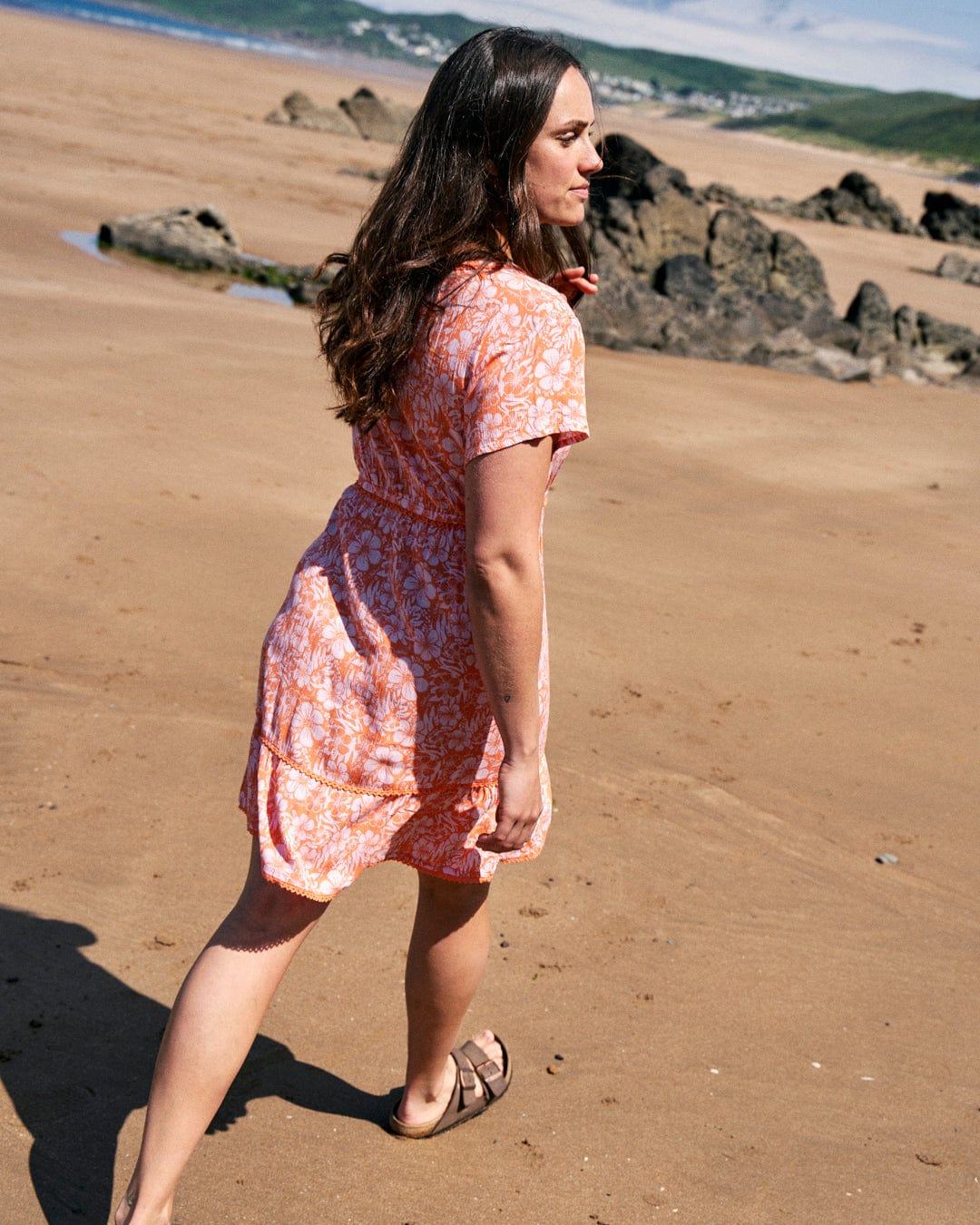 A woman in a Marla Hibiscus - Womens Dress - Orange by Saltrock walks barefoot on a sandy beach. Rocks and distant greenery are visible in the background.