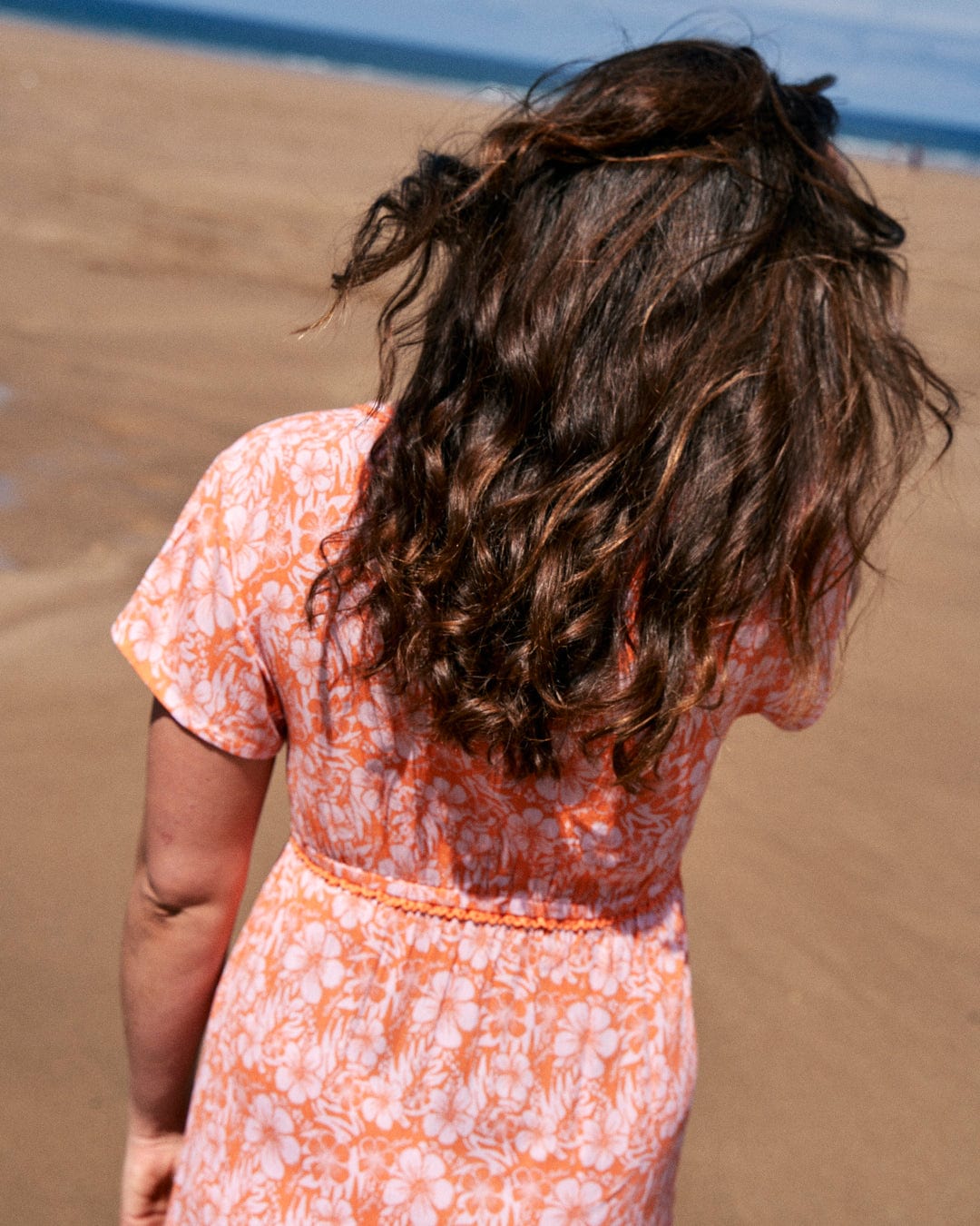 A woman with long, wavy brown hair stands on a sandy beach, wearing a Saltrock Marla Hibiscus - Womens Dress - Orange. She is facing away from the camera.