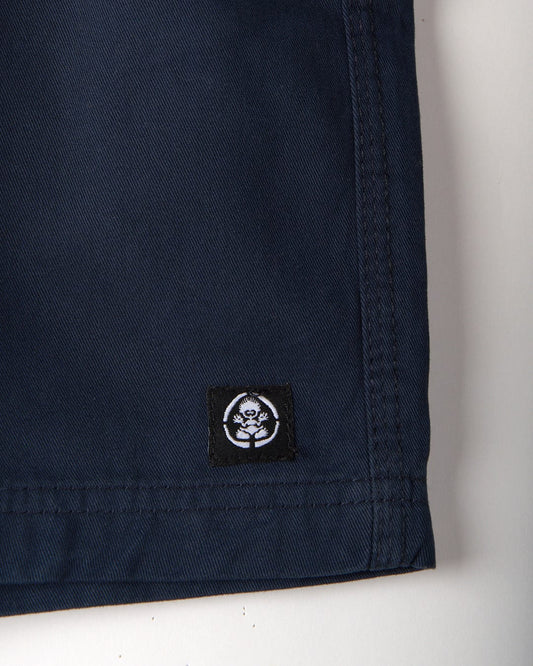 Close-up of the Meddon Kids Chino Shorts by Saltrock in dark blue cotton twill fabric, featuring visible stitching and a small black label with a white skull design.