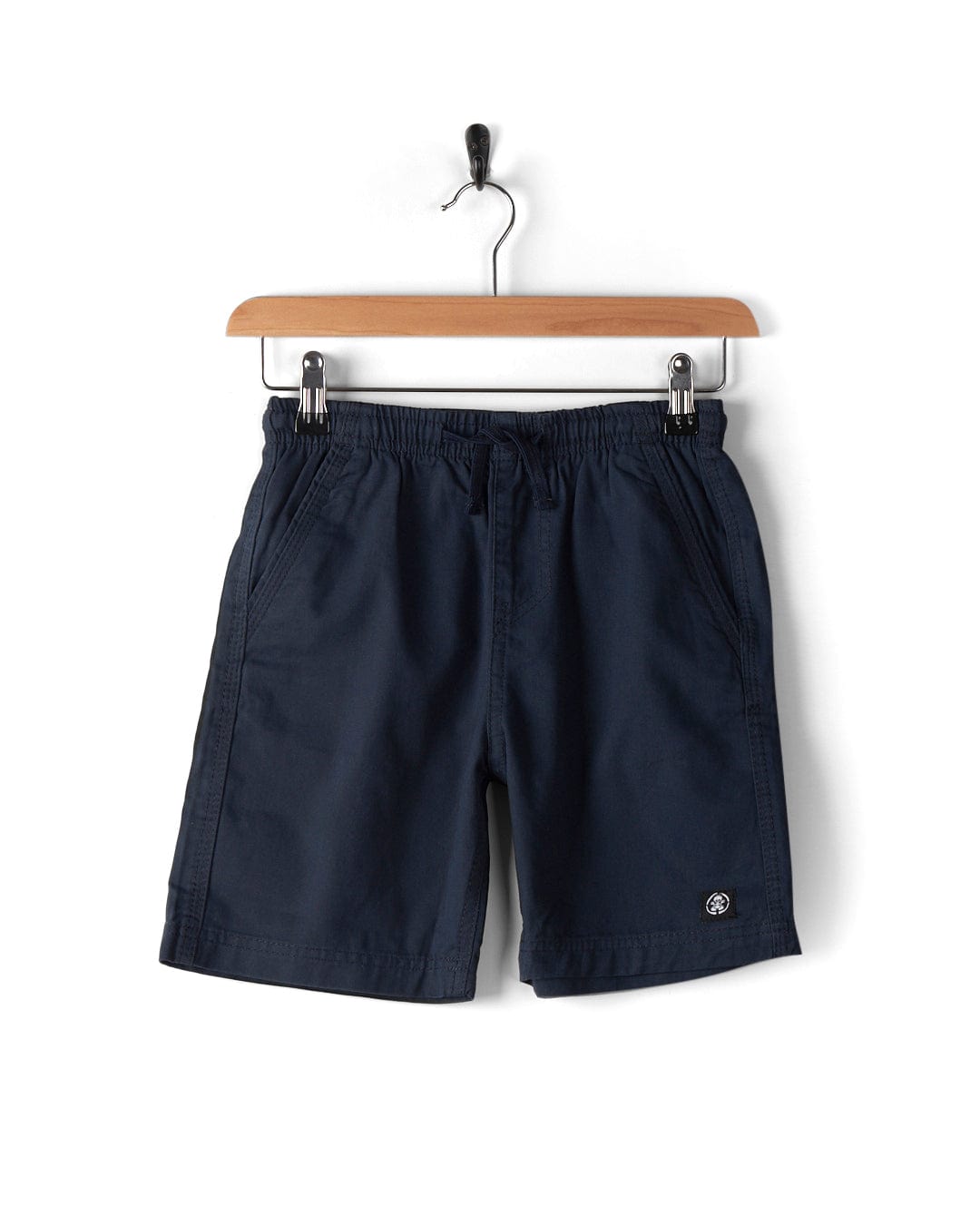 The Meddon - Kids Chino Shorts by Saltrock, in a classic navy blue color, feature a cotton twill fabric with an elasticated waistband and drawstring. They are displayed on a wooden hanger against a white background and are machine washable for easy care.