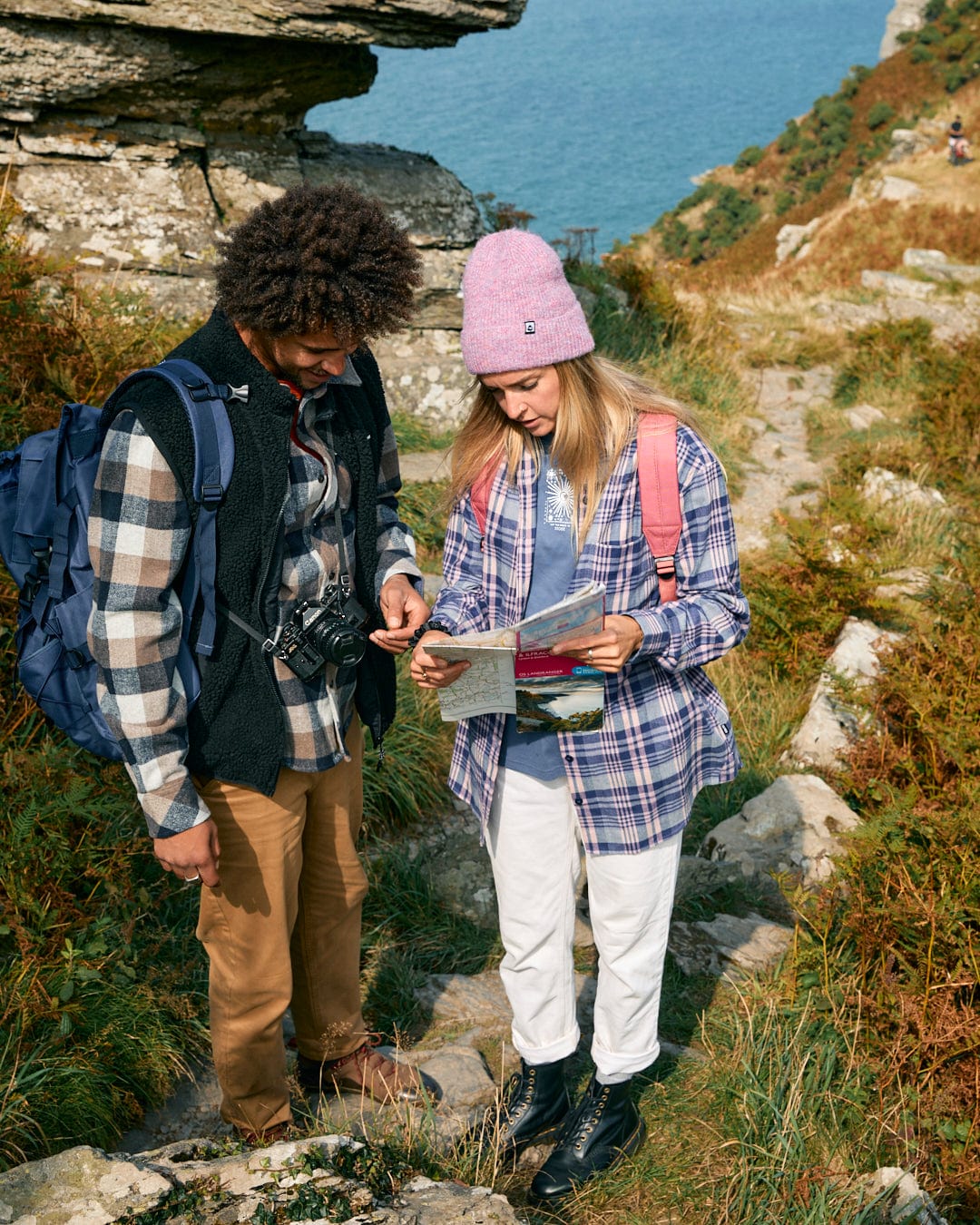 Two people stand on a rocky trail by the sea, checking their map. Both carry backpacks, wearing stylish Saltrock Kizzie women's longline shirts in purple and flannel beanies made from 100% cotton. The scene breathes adventure and timeless charm amidst the coastal breeze.