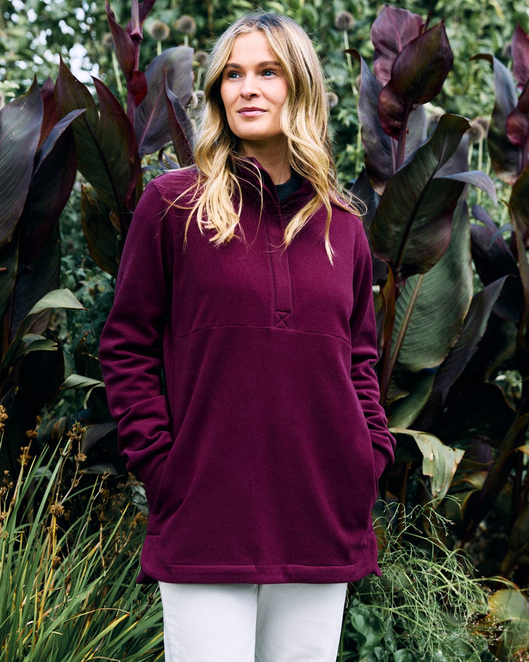 A person wearing the Morgan Women's 1/4 Neck Longline Fleece in Burgundy by Saltrock stands in a garden surrounded by large green leaves and plants.