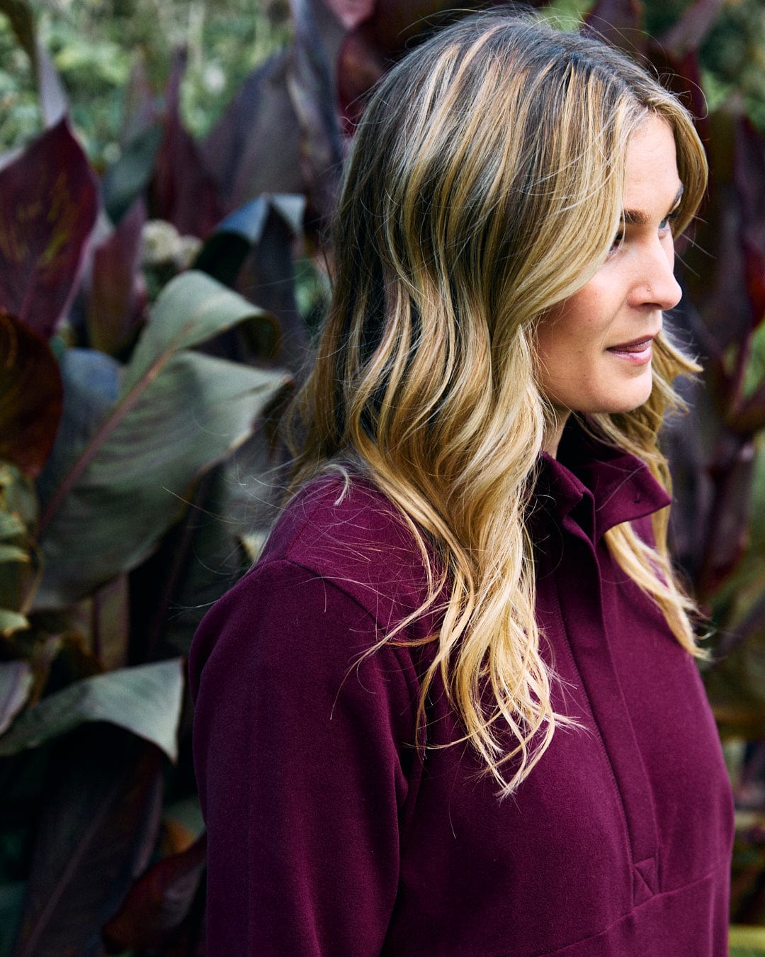 A person with long, wavy blonde hair stands in front of large, dark green leaves, wearing the Saltrock "Morgan" women's 1/4 neck longline fleece in burgundy.