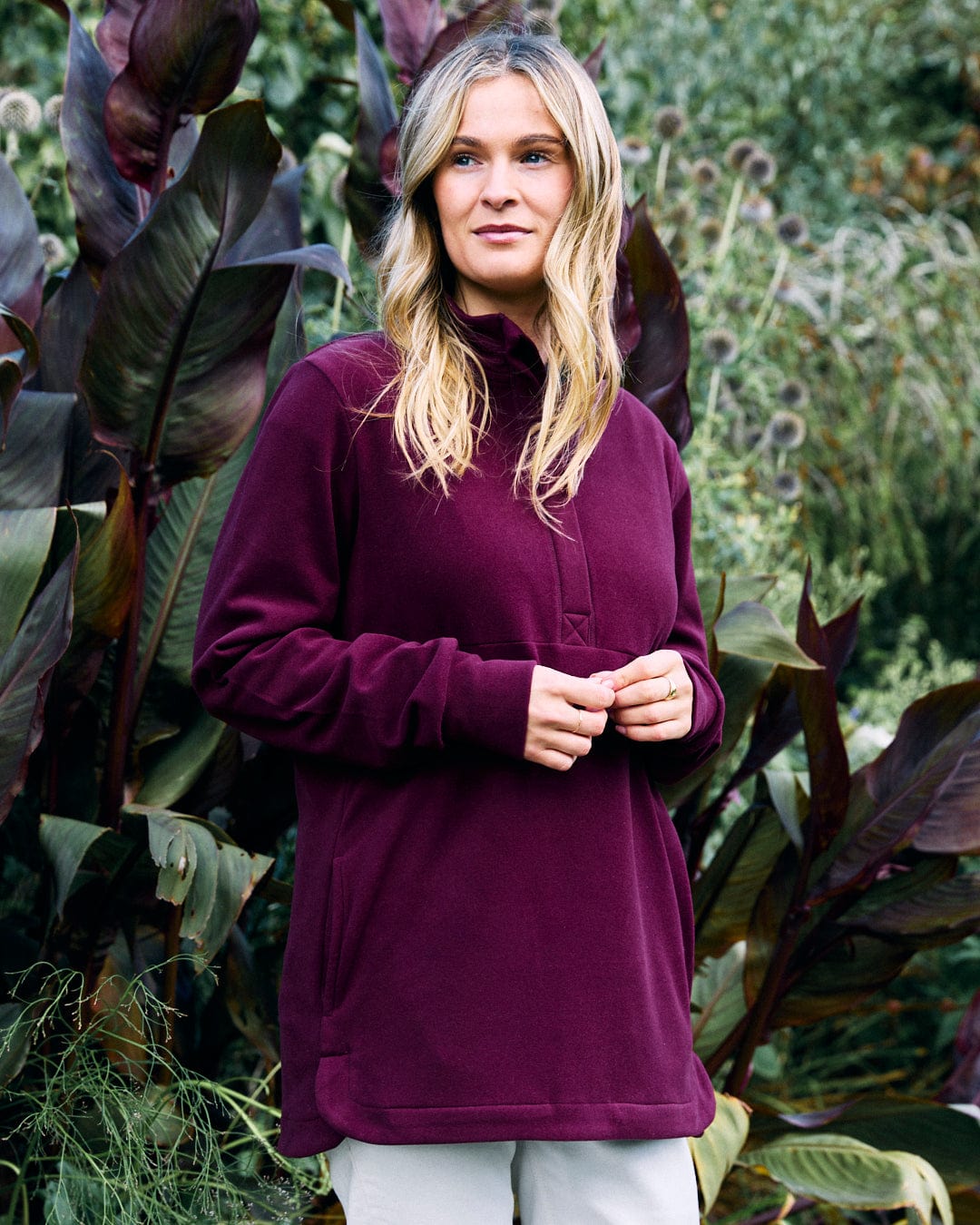 A person with long hair stands outdoors, wearing the Morgan Women’s 1/4 Neck Longline Fleece in burgundy by Saltrock, surrounded by lush green plants.