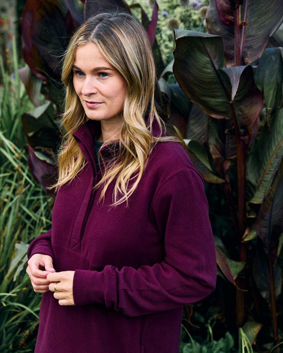 A person wearing the Saltrock Morgan Women's 1/4 Neck Longline Fleece in burgundy, with flowing hair, stands in front of large, leafy plants.