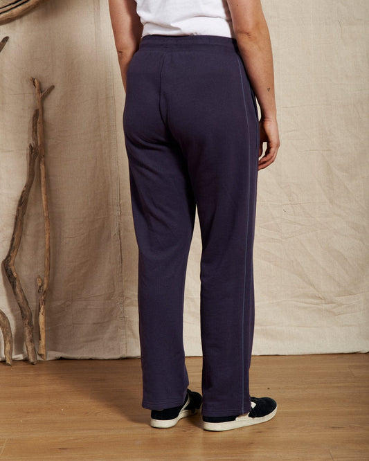 A person in purple Saltrock Morris joggers with a ribbed drawcord waist and black shoes stands on a wooden floor before a beige backdrop, with wooden sticks leaning against it. The cozy sweat material ensures comfort, while subtle embroidered branding adds style.