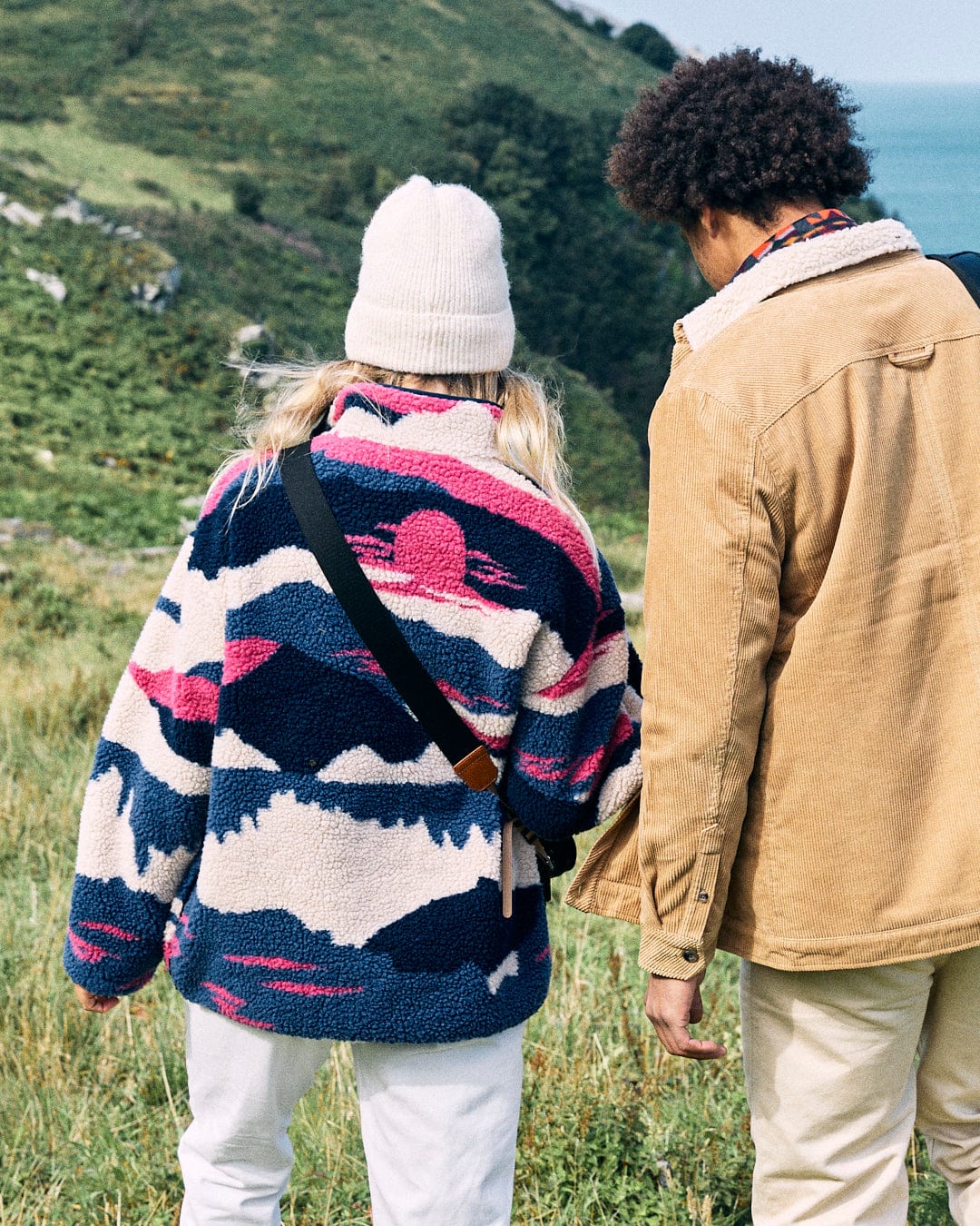 Two people in casual outdoor clothing stand on a grassy hillside, both wearing the Mountain Trail - Women's Recycled Borg Zip Fleece by Saltrock. The jackets, featuring a subtle mountain landscape print and sherpa-lined collars, keep them cozy as they gaze at the ocean view in the background.