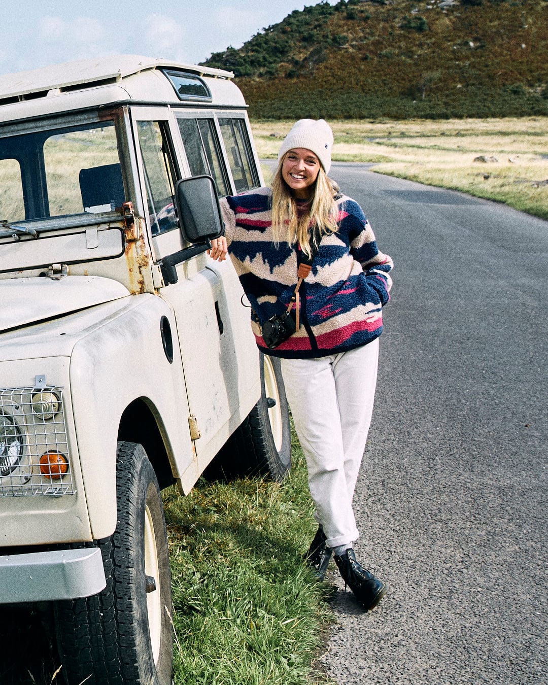 A person in a blue Saltrock Mountain Trail Women's Recycled Sherpa Zip Fleece and white pants leans against an off-road vehicle parked on a paved road with grassy hills in the background.