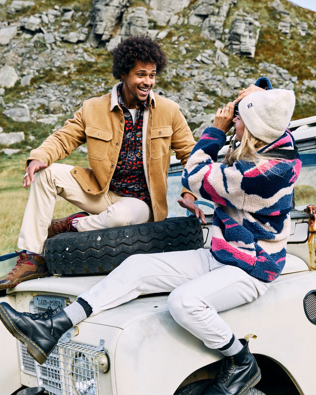 Two people sit on a white Land Rover in an outdoor, rocky area. One person is wearing a brown jacket, and the other is wearing the Saltrock Mountain Trail - Women's Recycled Sherpa Zip Fleece in blue. They smile and engage with each other, enjoying the moment.