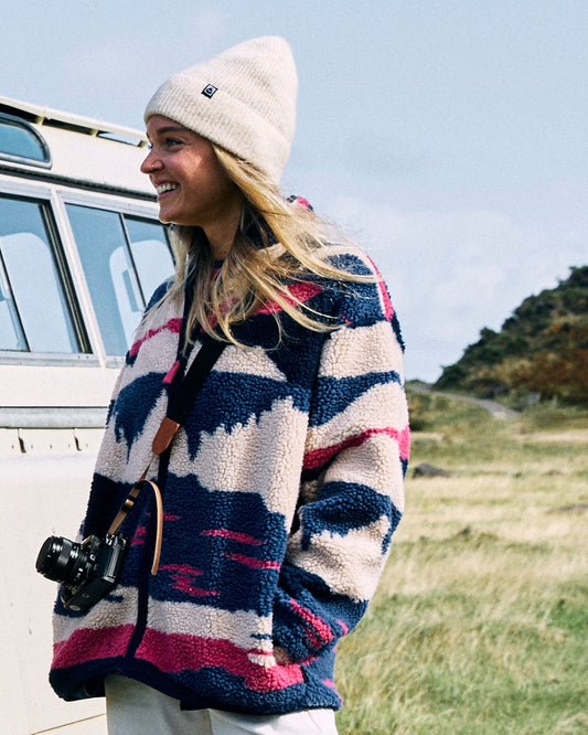 A woman wearing a Saltrock Mountain Trail Women's Recycled Sherpa Zip Fleece in blue and a beanie stands outside by a vehicle, holding a camera and smiling.