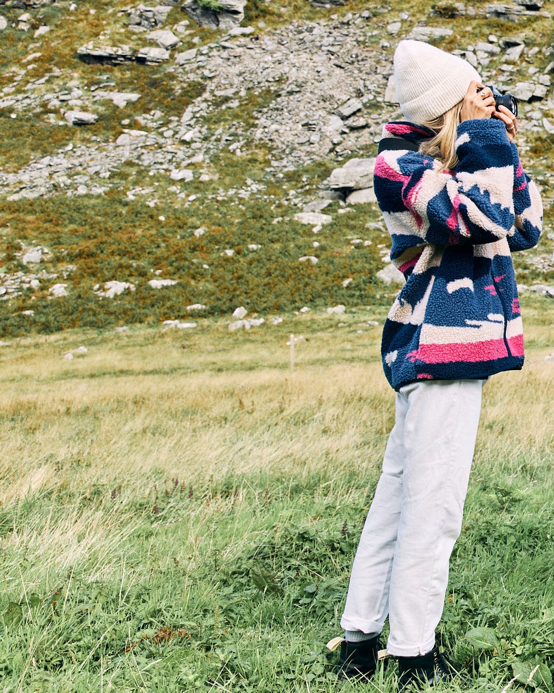 A person wearing a Saltrock Mountain Trail Women's Recycled Sherpa Zip Fleece in blue, white pants, and a beanie stands in a grassy area taking a photo with a camera. The machine-washable jacket features a full zip opening. A rocky hillside is in the background.