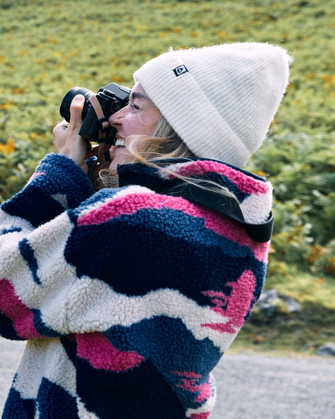 The person, draped in a blue Saltrock Mountain Trail Women's Recycled Sherpa Zip Fleece, and sporting a white beanie, is capturing a photo in an outdoor setting surrounded by lush greenery.