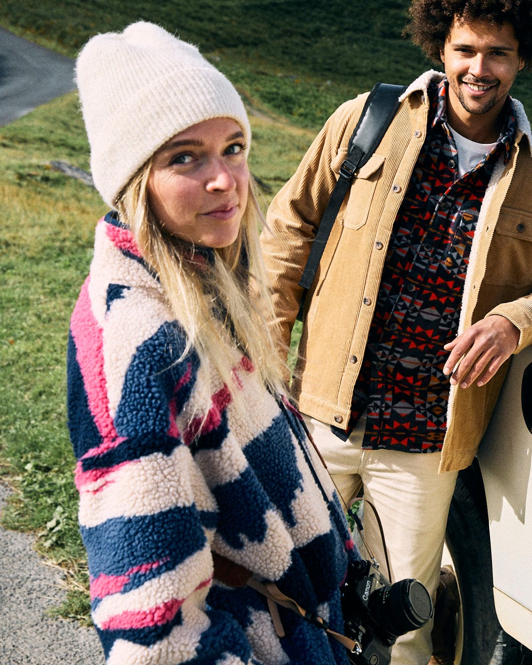 Two people are outdoors; the woman is wearing the Saltrock Mountain Trail - Womens Recycled Sherpa Zip Fleece in blue, along with a white hat, holding a camera. The man is donning a machine washable jacket, plaid shirt, and backpack. Both are smiling while standing in a grassy area.