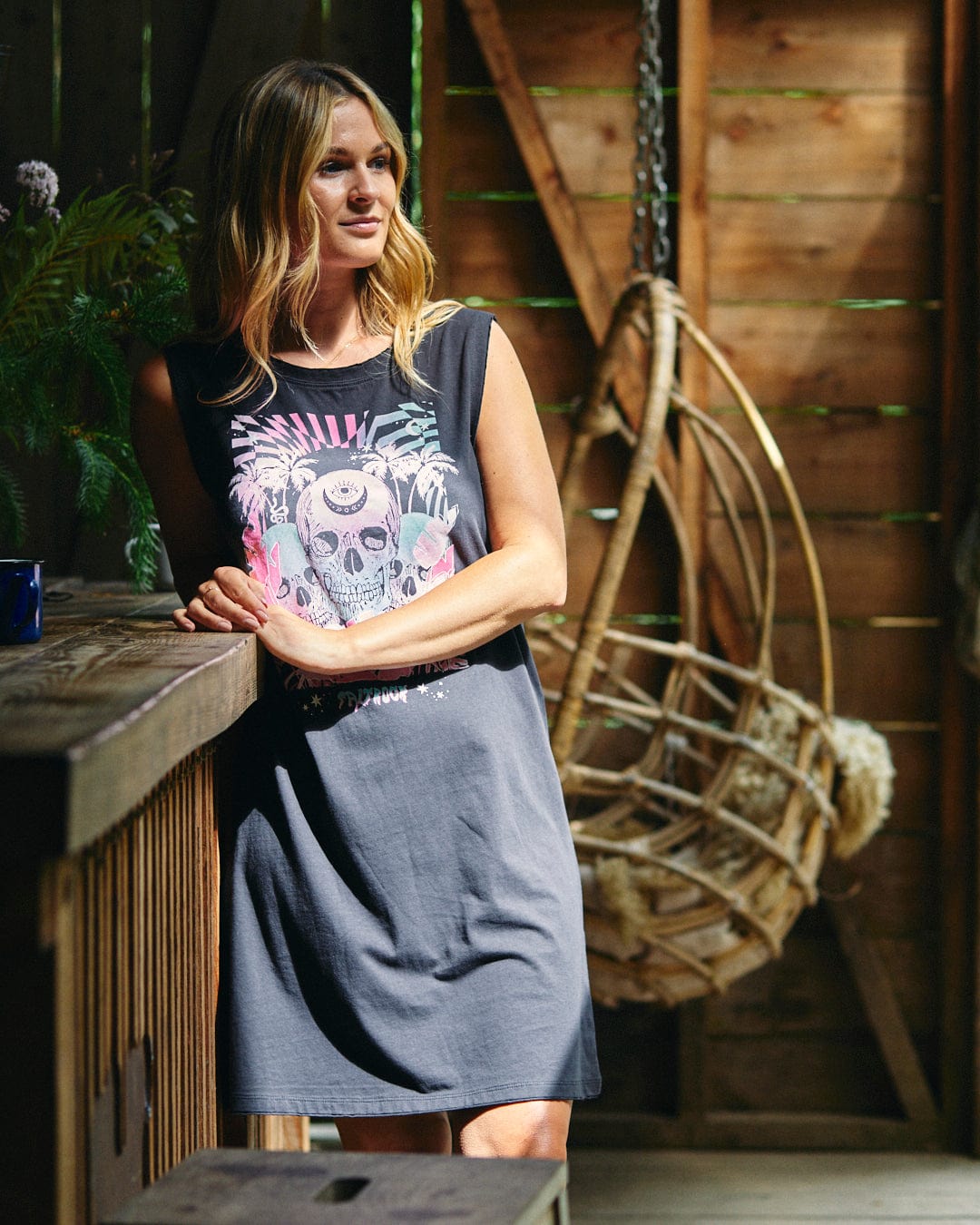 A woman wearing the Saltrock Mystic Skulls - Women's Jersey Vest Midi Dress in black, characterized by a neon warped skull illustration stands by a rustic wooden counter. Made of 100% cotton, this exclusive dress also features Saltrock branding. A hanging wicker chair is visible in the background.