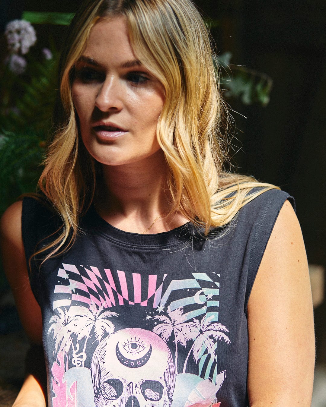 A person with long, blonde hair is wearing the Mystic Skulls - Women's Jersey Vest Midi Dress from Saltrock, which is made of 100% cotton and features a colorful graphic design with a neon warped skull illustration alongside palm trees. They are looking slightly to the side in a dimly lit environment.