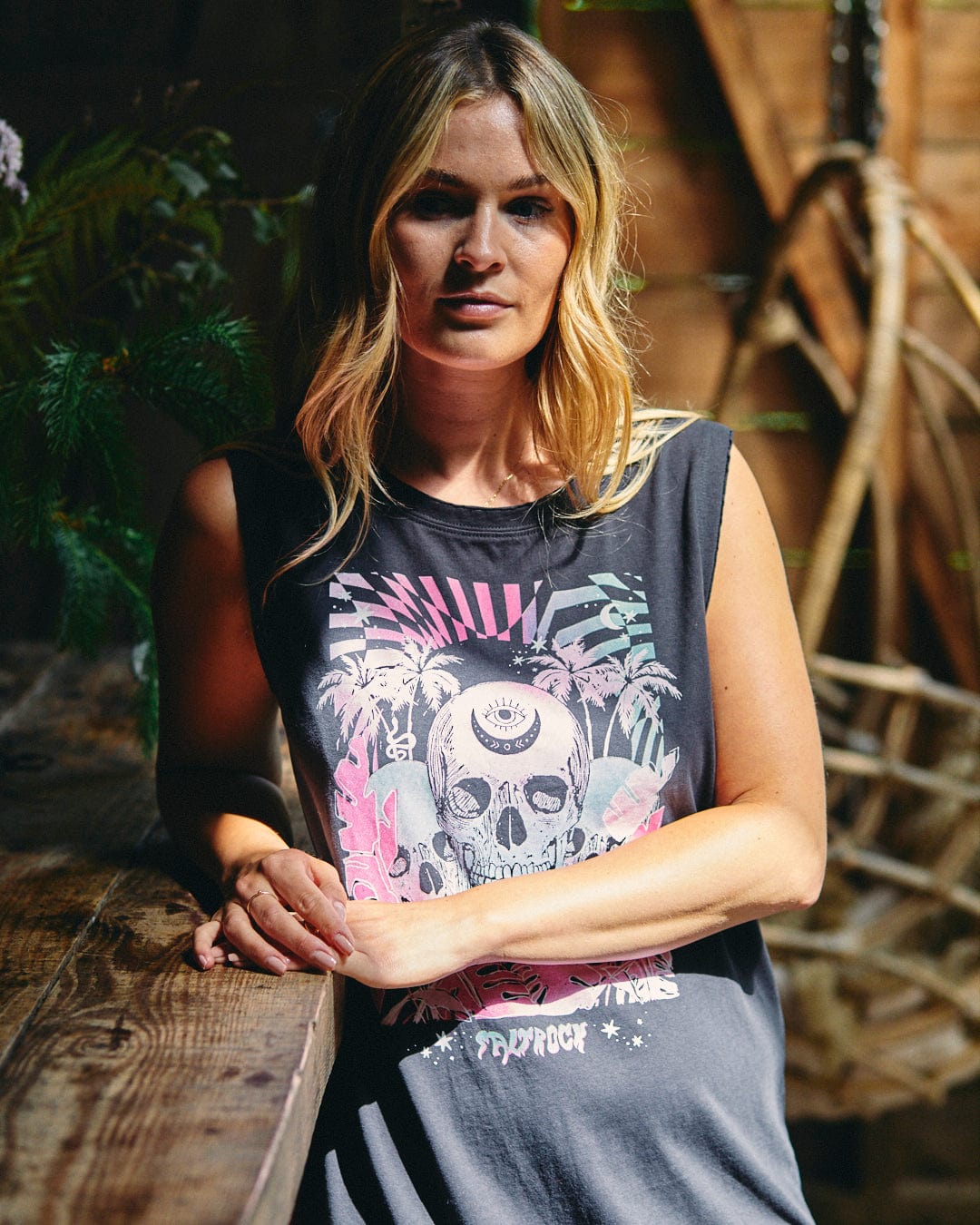 A woman with blonde hair stands next to a wooden table in a room with rustic decor, wearing the Saltrock Mystic Skulls Women's Jersey Vest Midi Dress, which features a neon warped skull illustration.