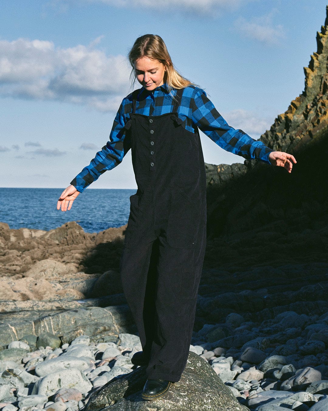 A fashion-conscious woman in a **Nancy - Womens Cord Jumpsuit - Dark Blue** by **Saltrock** stands on a rocky shoreline, balancing with arms extended, with the ocean and cliffs in the background.