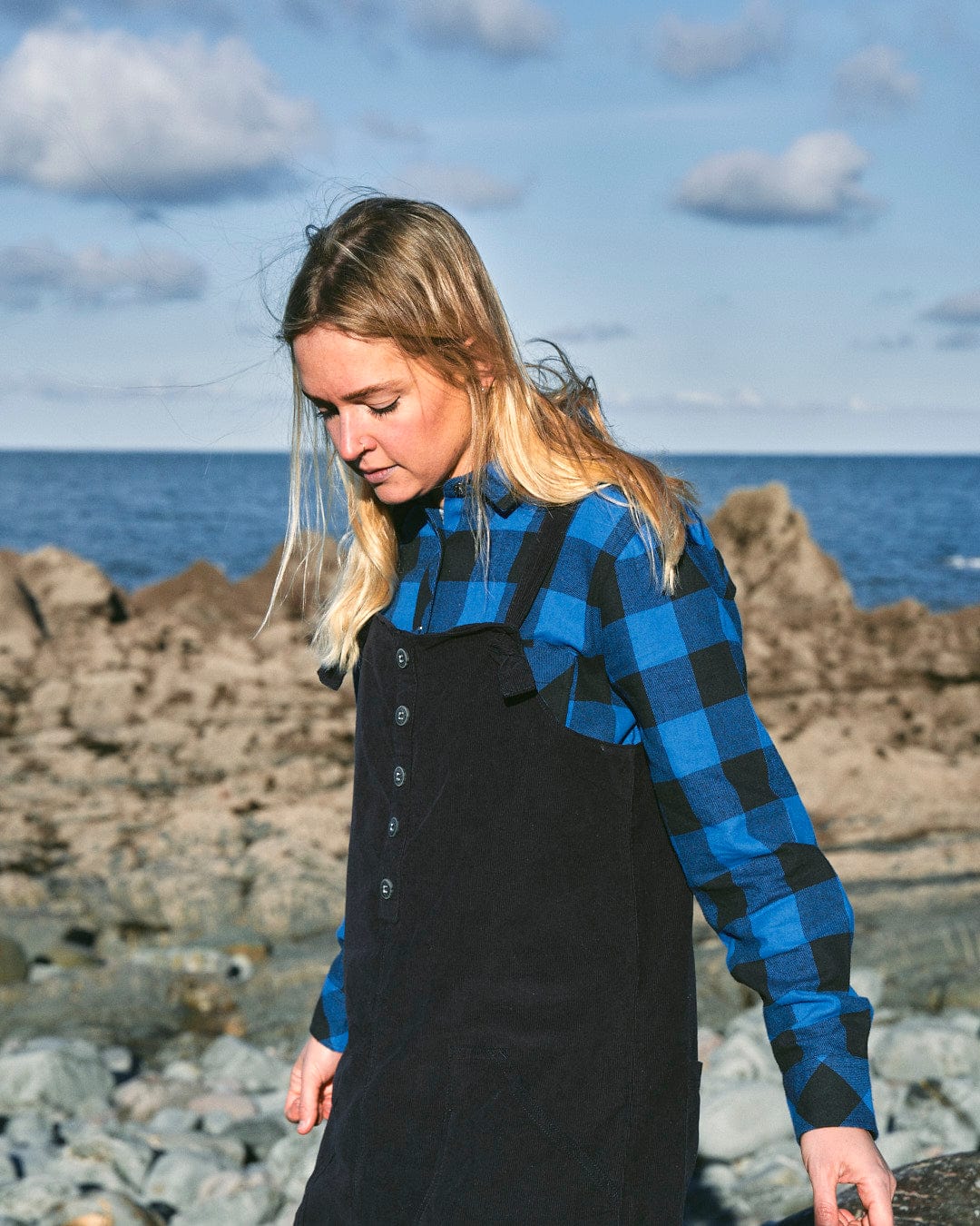 A fashion-conscious woman with long blonde hair, wearing a Nancy - Womens Cord Jumpsuit - Dark Blue by Saltrock and a blue plaid shirt, walks on a rocky beach with the ocean and a partly cloudy sky in the background.