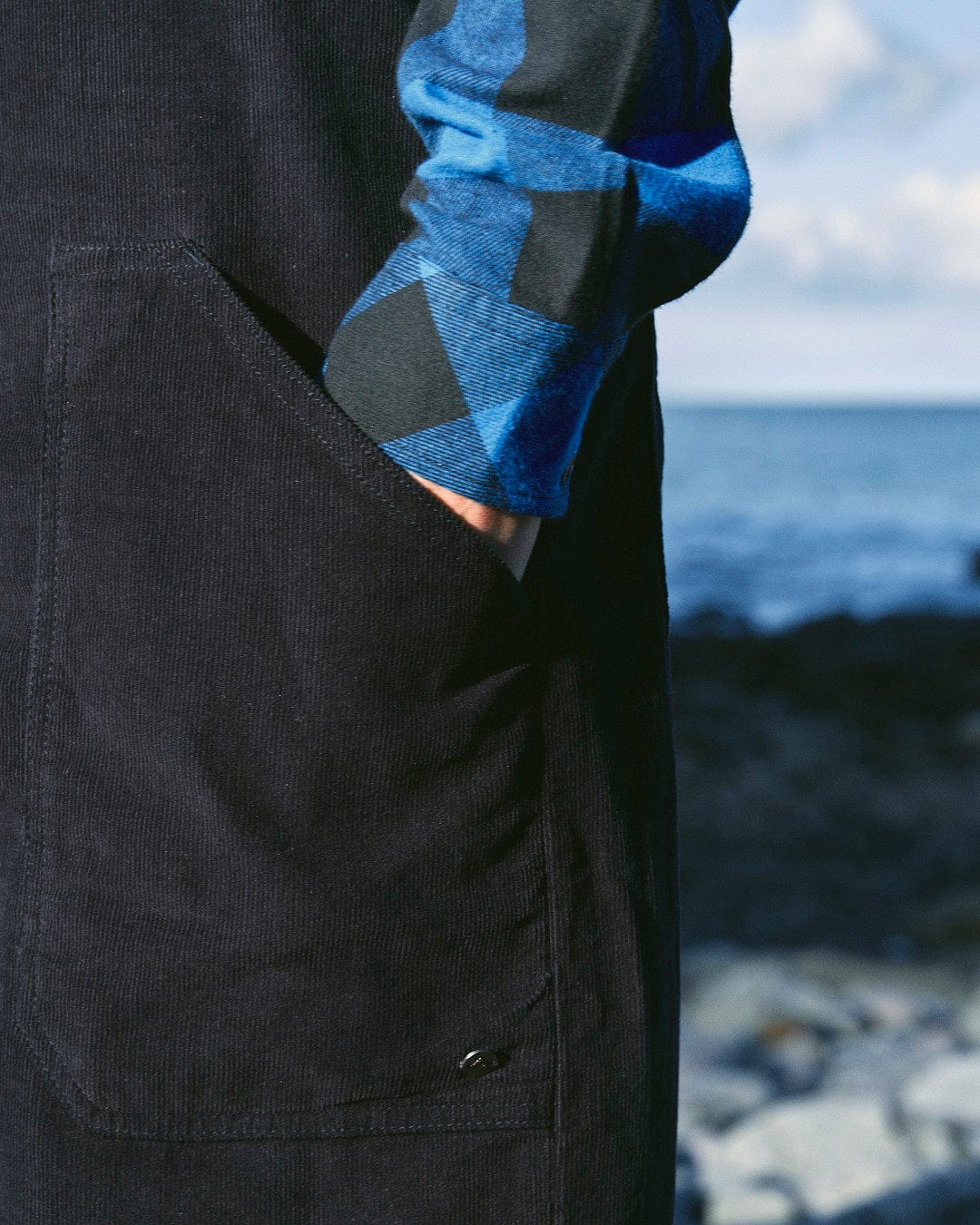 Close-up of a fashion-conscious woman wearing a blue and black checkered shirt with one hand in the pocket of her Saltrock Nancy - Womens Cord Jumpsuit - Dark Blue, standing near a rocky shoreline with the ocean in the background.