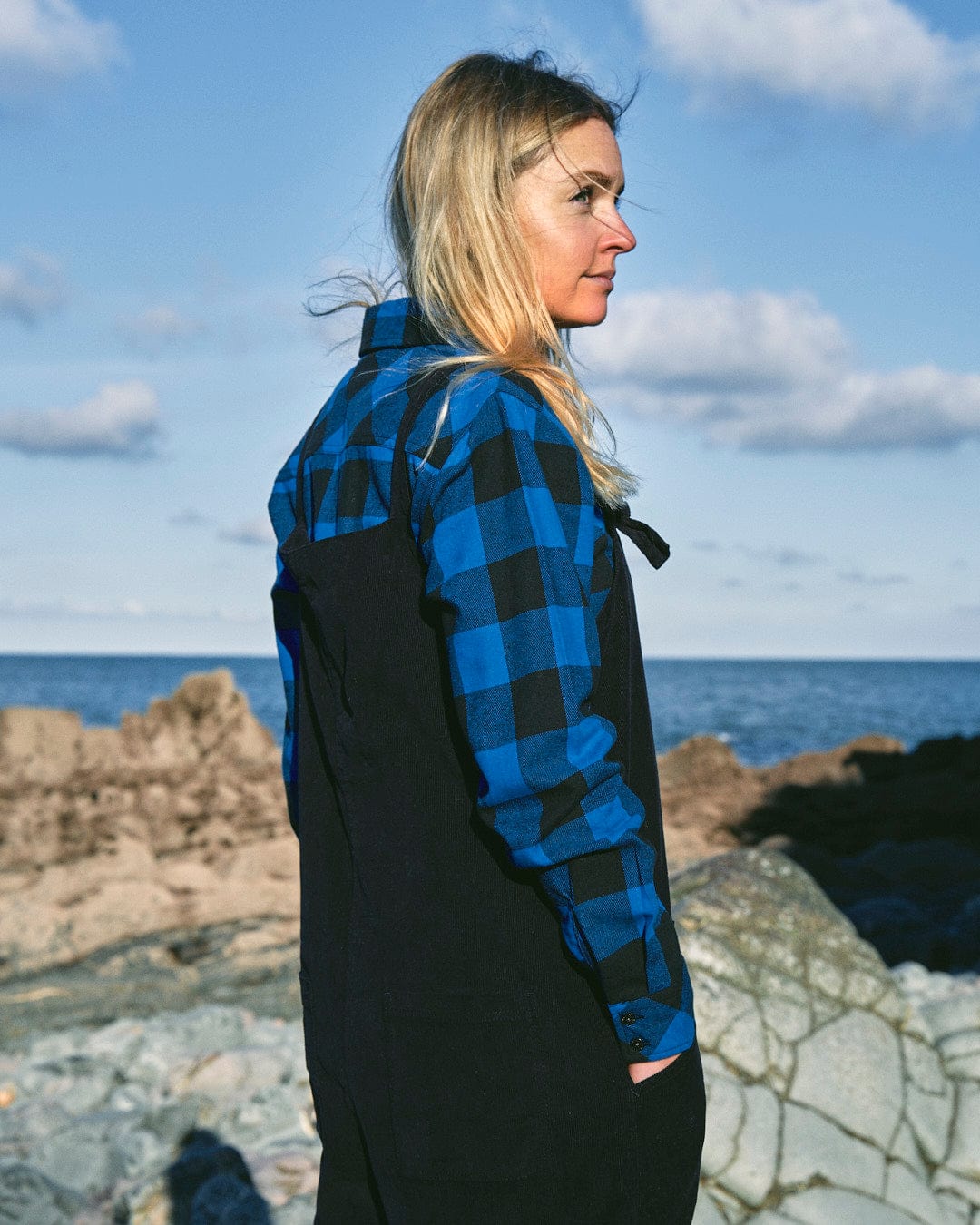 A fashion-conscious woman with long blonde hair, dressed in a Saltrock Nancy - Womens Cord Jumpsuit - Dark Blue and black overalls, stands on rocky terrain near the ocean, looking into the distance.