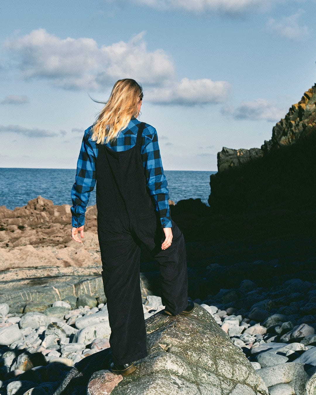 A fashion-conscious woman with long hair, wearing a blue checkered shirt and Saltrock's Nancy - Womens Cord Jumpsuit - Dark Blue, stands on a rocky coastline looking out towards the sea.