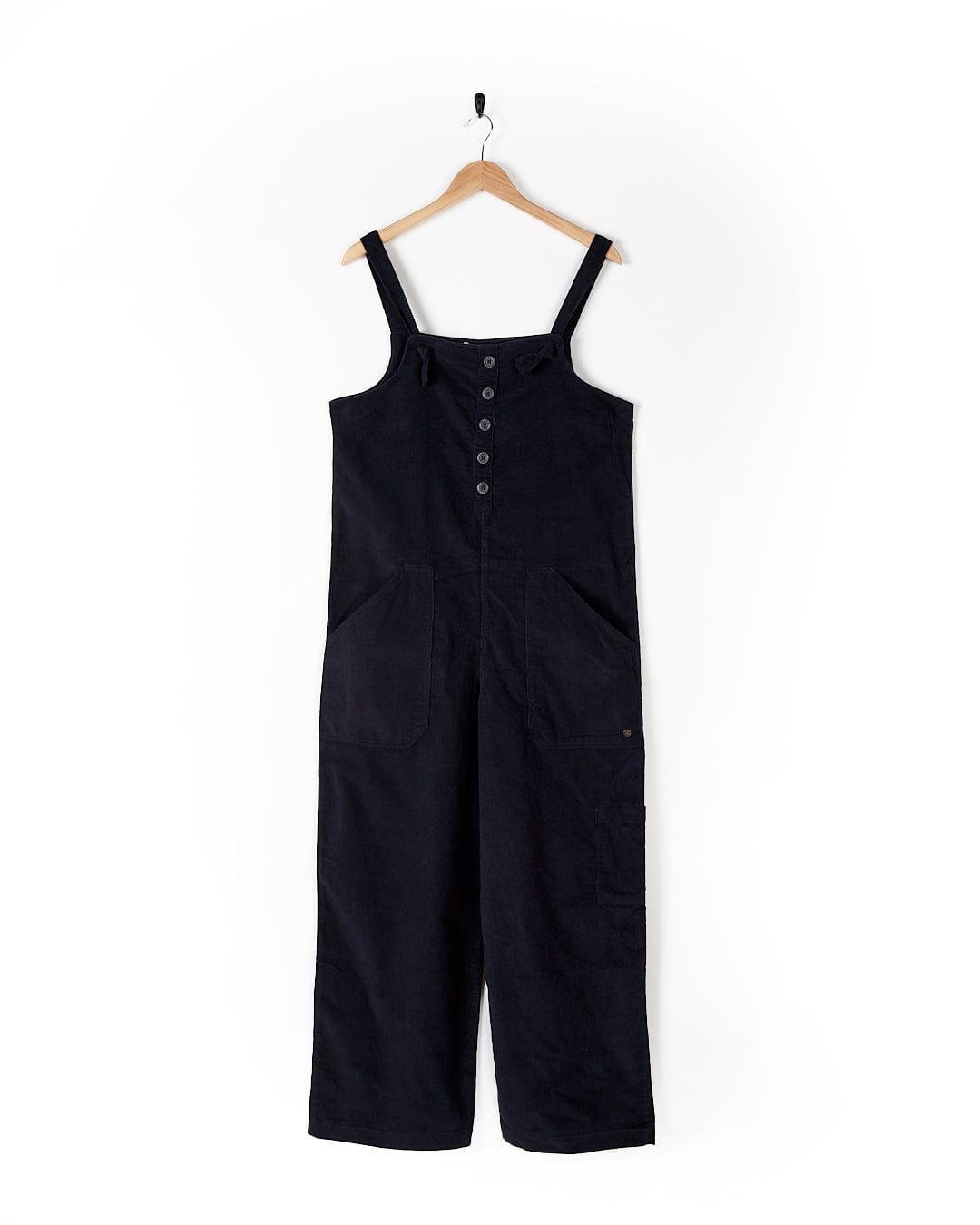 A Saltrock Nancy - Womens Cord Jumpsuit - Dark Blue is hanging on a wooden hanger against a white background. Fashion-conscious women will appreciate the wide legs, button details on the chest, and two large front pockets, blending both style and practicality effortlessly.