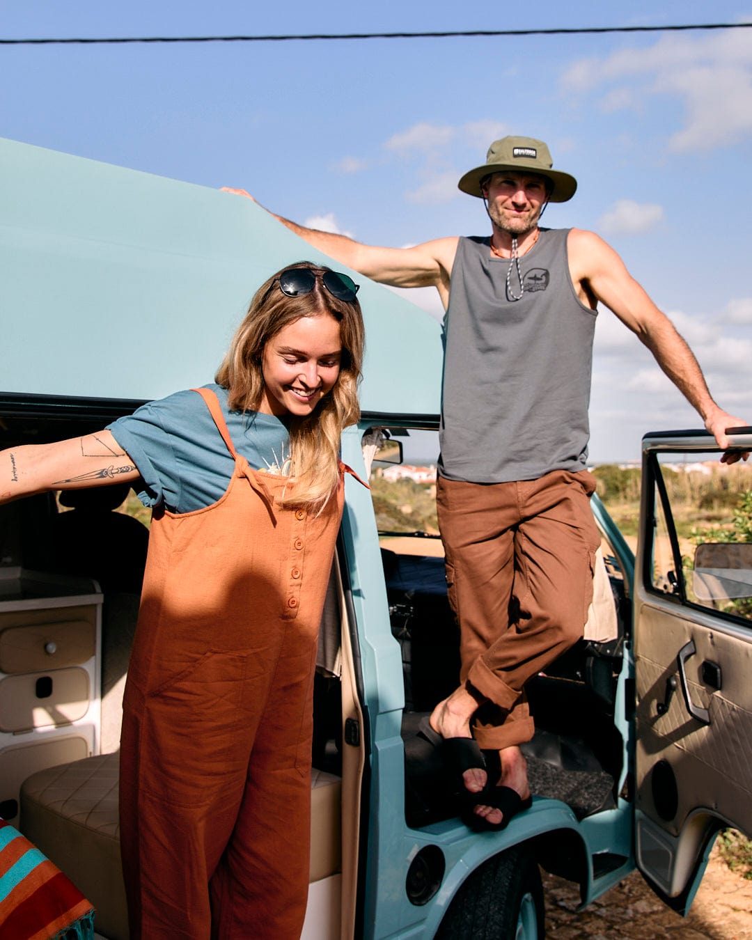 Two people stand outside of a light blue van. The woman on the left wears Saltrock Nancy - Womens Dungarees - Brown with adjustable tie straps and a teal shirt, while the man on the right, in a relaxed fit gray tank top and brown pants, casually tips his hat.