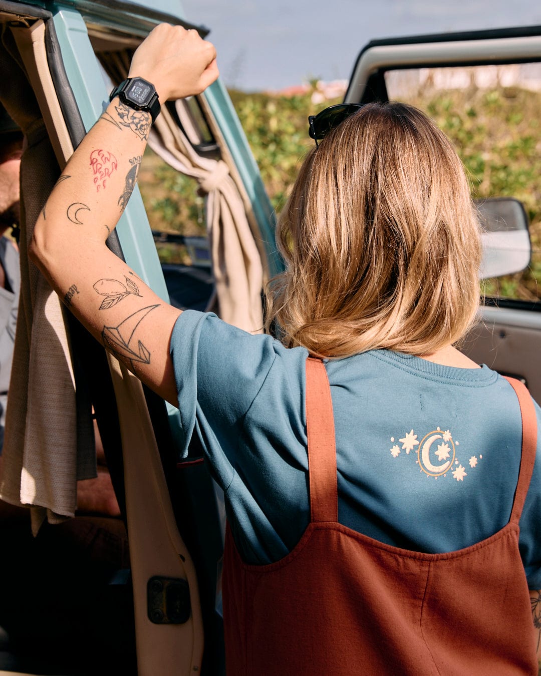 Person with tattoos on their arms stands beside an open van door, facing away from the camera, wearing a blue shirt with a celestial design and Saltrock Nancy - Womens Dungarees - Brown featuring adjustable tie straps.