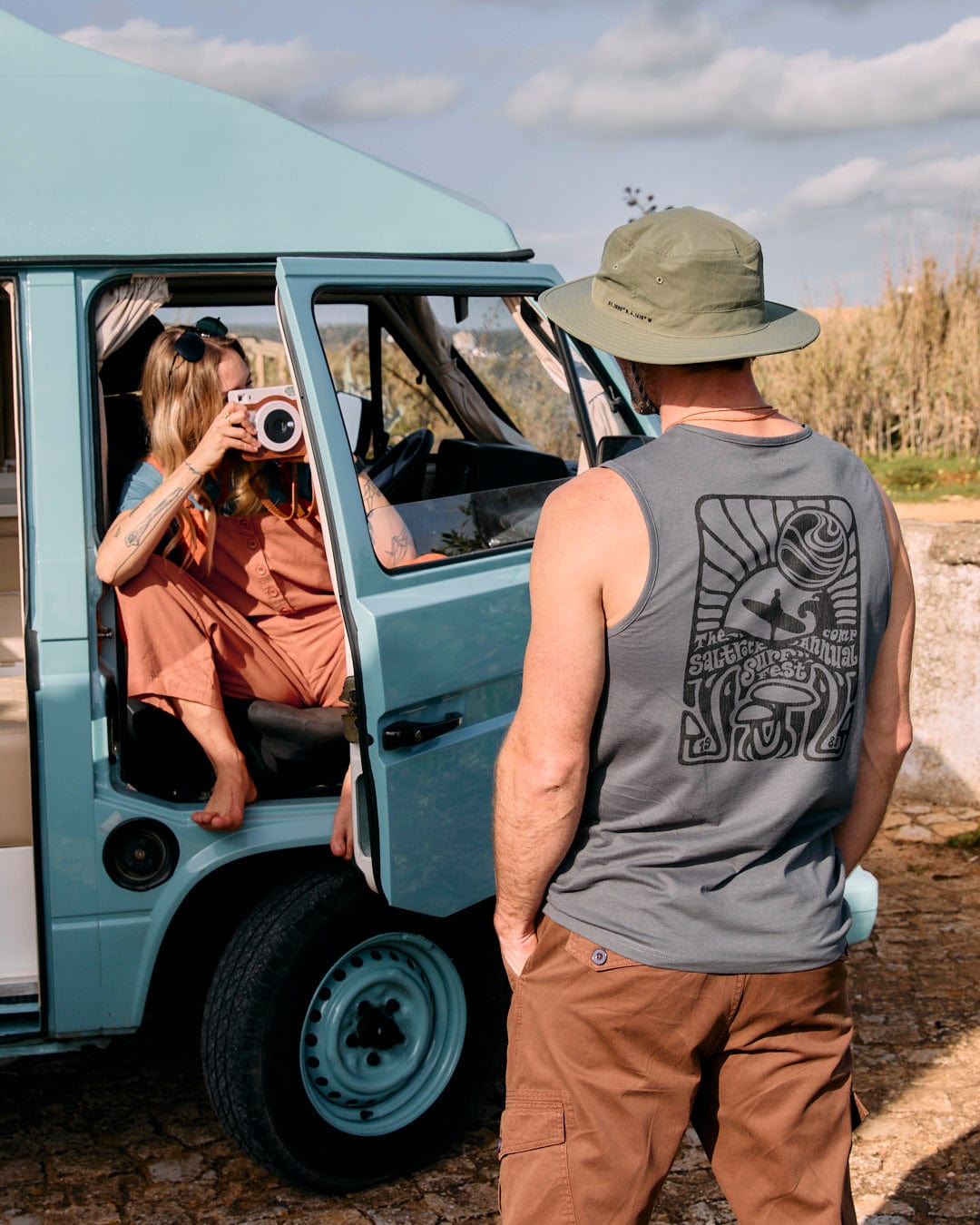 Person in a sleeveless shirt and hat stands outside a teal-colored van while another person inside the van, dressed in Saltrock Nancy - Womens Dungarees - Brown, takes a photo with an instant camera.