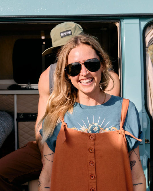 A smiling woman in a Nancy - Womens Jumpsuit - Brown by Saltrock stands in front of a teal van, her sunglasses reflecting the sunlight. Another person wearing a green cap, partly hidden behind her, peeks out shyly.