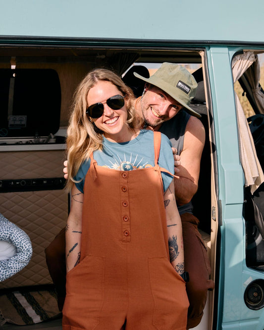 A woman in a relaxed fit Nancy - Womens Jumpsuit - Brown from Saltrock and a blue shirt smiles, wearing sunglasses, with a man in a green hat leaning from behind her. They are standing near the open door of a retro-style van.