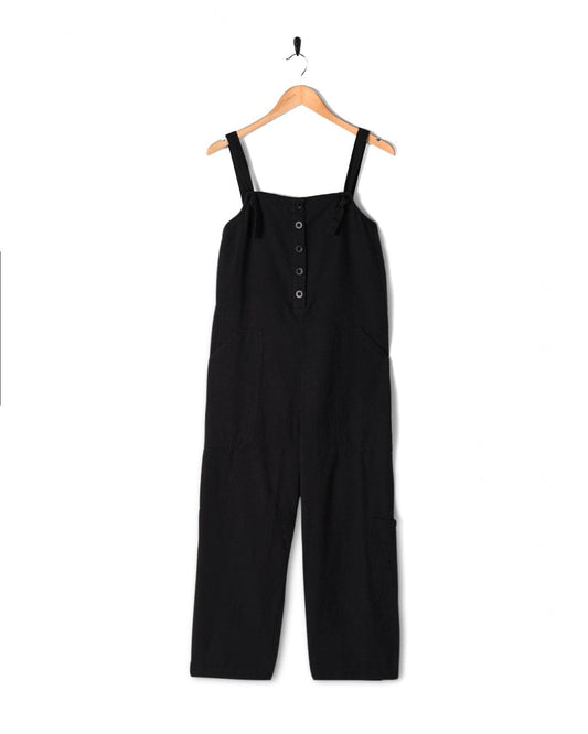 A Nancy - Womens Jumpsuit - Dark Grey with button details down the front and adjustable tie straps, made from soft cotton linen, displayed on a wooden hanger against a white background by Saltrock.