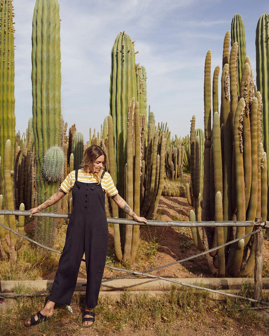 A person in a relaxed fit, striped shirt and wearing the Saltrock Nancy - Womens Jumpsuit - Dark Grey stands by a metal fence in front of a dense field of tall cacti.