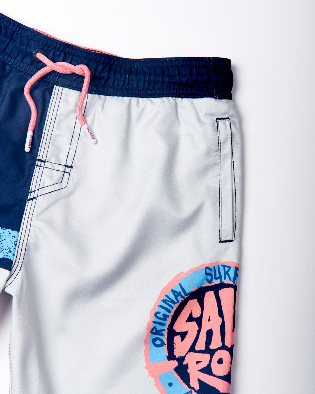 Image of light-colored swim trunks with a blue waistband and a pink drawstring featuring branded dipped rubber ends. The Saltrock Original SR - Kids Swimshorts - Blue have an elasticated waist and a pink and blue graphic displaying the words "Original Surf" and "SALE" near the hem, along with subtle Saltrock branding.